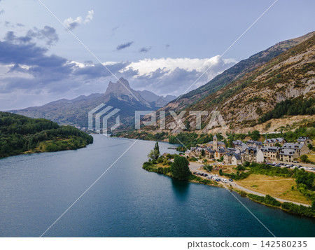 Mountain Village and Reservoir Below Foratata. Aerial view of a mountain village beside the turquoise Lanuza reservoir with the Pena Foratata peak on the horizon near Formigal, Spain, a serene Pyrenee Mountain Village and Reservoir Below Foratata. Aerial view of a mountain village beside the turquoise Lanuza reservoir with the Pena Foratata peak on the horizon near Formigal, Spain, a serene Pyrenee 142580235