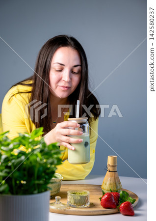 Smiling Woman Sipping Matcha Latte with Straw, Summer. Cheerful woman sipping green matcha latte through straw with content smile in cozy summer setting. Rustic wooden background with ceramic bowl Smiling Woman Sipping Matcha Latte with Straw, Summer. Cheerful woman sipping green matcha latte through straw with content smile in cozy summer setting. Rustic wooden background with ceramic bowl 142580237