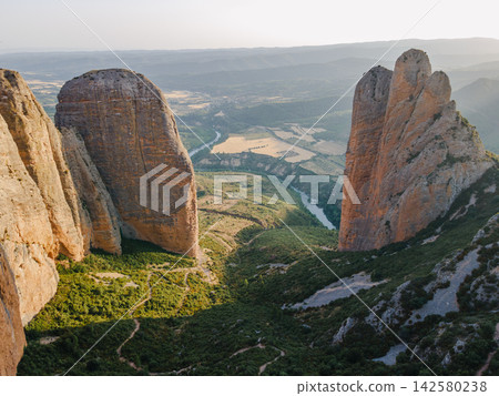 Towering Red Rock Walls of Mallos de Riglos. Aerial view of the towering red rock walls of the Mallos de Riglos rising above the green valley in Aragon, Spain, a majestic natural landmark and a famous Towering Red Rock Walls of Mallos de Riglos. Aerial view of the towering red rock walls of the Mallos de Riglos rising above the green valley in Aragon, Spain, a majestic natural landmark and a famous 142580238