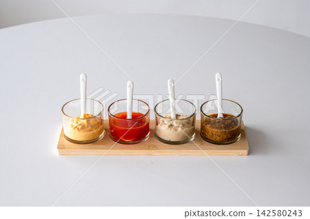 Mustard and dipping sauces row on bamboo board side view. Side view of mustard and dipping sauces arranged in row on bamboo board with white spoons, gourmet condiment lineup for BBQ grilling and food Mustard and dipping sauces row on bamboo board side view. Side view of mustard and dipping sauces arranged in row on bamboo board with white spoons, gourmet condiment lineup for BBQ grilling and food 142580243
