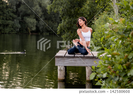 Young Woman in Seated Cross Legged Pose On A Dock in the Park at Sunset. A young Hispanic woman practises a seated cross legged pose on a dock surrounded by greenery in a park at golden hour, a moment 142580608