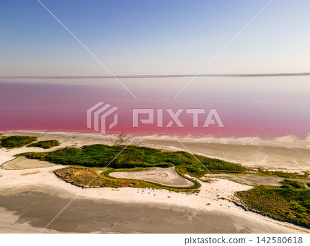 Green Island on the Pink Salt Flats. Aerial view of a green island resting on the pink salt flats of Lake Sivash beneath a pale blue horizon in Ukraine, the soft colours of an unusual natural pink lak Green Island on the Pink Salt Flats. Aerial view of a green island resting on the pink salt flats of Lake Sivash beneath a pale blue horizon in Ukraine, the soft colours of an unusual natural pink lak 142580618