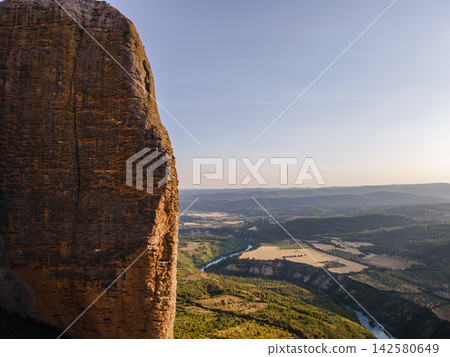 Rock Face of Mallos de Riglos Above the Valley. Aerial view of a towering conglomerate rock face of the Mallos de Riglos above the Gallego river valley in Aragon, Spain, a majestic natural landmark Rock Face of Mallos de Riglos Above the Valley. Aerial view of a towering conglomerate rock face of the Mallos de Riglos above the Gallego river valley in Aragon, Spain, a majestic natural landmark 142580649