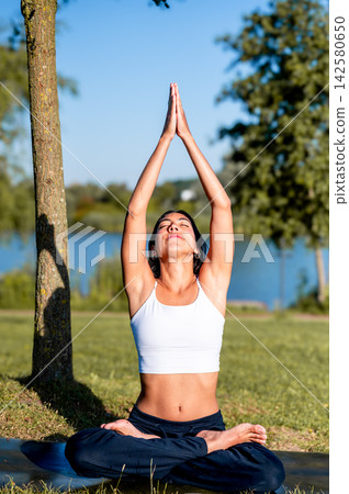 Hispanic Woman in Seated Pose With Arms Overhead on a Dock by the Lake. A young Hispanic woman practises a seated pose with arms overhead on a wooden dock over the calm lake at sunset, a practice Hispanic Woman in Seated Pose With Arms Overhead on a Dock by the Lake. A young Hispanic woman practises a seated pose with arms overhead on a wooden dock over the calm lake at sunset, a practice 142580650