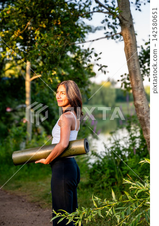 Hispanic Woman in Woman Carrying a Yoga Mat in a Park. A young Hispanic woman carries a rolled yoga mat along a park path by the lake at golden hour on her way to an outdoor yoga session, embracing Hispanic Woman in Woman Carrying a Yoga Mat in a Park. A young Hispanic woman carries a rolled yoga mat along a park path by the lake at golden hour on her way to an outdoor yoga session, embracing 142580651