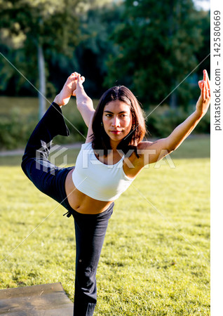 Fit Woman in Extended Hand To Big Toe Pose by the Lakeside at Golden Hour. A young Hispanic woman practises the extended hand to big toe pose in a peaceful park by the lakeside in summer, for relaxati Fit Woman in Extended Hand To Big Toe Pose by the Lakeside at Golden Hour. A young Hispanic woman practises the extended hand to big toe pose in a peaceful park by the lakeside in summer, for relaxati 142580669