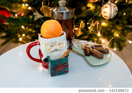 Santa Mug with Orange and Cinnamon by Christmas Tree. A festive Santa Claus mug holding an orange beside a glass jar of tea and a plate of cinnamon sticks and chocolate on a white table before a glow Santa Mug with Orange and Cinnamon by Christmas Tree. A festive Santa Claus mug holding an orange beside a glass jar of tea and a plate of cinnamon sticks and chocolate on a white table before a glow 142580672