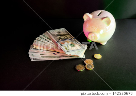 Dollar Bills Beside a Pink Piggy Bank. A fan of US dollar bills beside a pink piggy bank and coins on a dark background, a conceptual image of saving money, budgeting and personal finance. Dollar Bills Beside a Pink Piggy Bank. A fan of US dollar bills beside a pink piggy bank and coins on a dark background, a conceptual image of saving money, budgeting and personal finance. 142580673