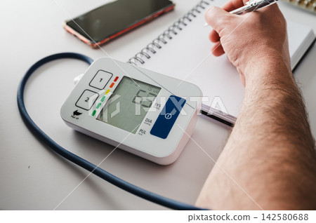 Close-up of Digital Blood Pressure Monitor and Notebook. Close-up of a digital home blood pressure monitor beside a smartphone and a notebook with a hand recording the reading, illustrating hypertensi Close-up of Digital Blood Pressure Monitor and Notebook. Close-up of a digital home blood pressure monitor beside a smartphone and a notebook with a hand recording the reading, illustrating hypertensi 142580688