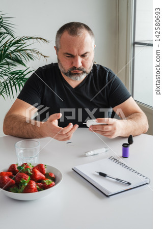 Mature Man Doing Blood Sugar Test with Fresh Strawberries. Mature man does a finger- blood sugar test with a glucometer at a table with a bowl of fresh strawberries and water, illustrating diabetes se Mature Man Doing Blood Sugar Test with Fresh Strawberries. Mature man does a finger- blood sugar test with a glucometer at a table with a bowl of fresh strawberries and water, illustrating diabetes se 142580693