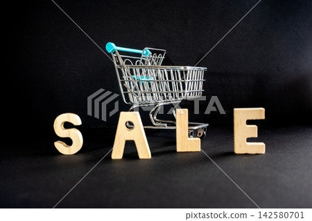 SALE Letters and a Miniature Shopping Cart. The word SALE spelled in wooden letters beside a miniature shopping cart on a dark background with colorful lighting, a creative concept of shopping, discou SALE Letters and a Miniature Shopping Cart. The word SALE spelled in wooden letters beside a miniature shopping cart on a dark background with colorful lighting, a creative concept of shopping, discou 142580701