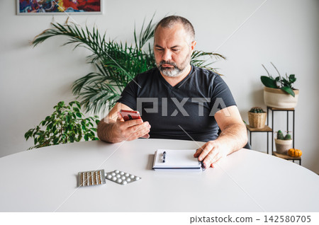 Mature Man Reading Medication Information at Home Table. A mature man reads medication information while holding pills at a table with blister packs, a notebook and a smartphone, illustrating medicati Mature Man Reading Medication Information at Home Table. A mature man reads medication information while holding pills at a table with blister packs, a notebook and a smartphone, illustrating medicati 142580705