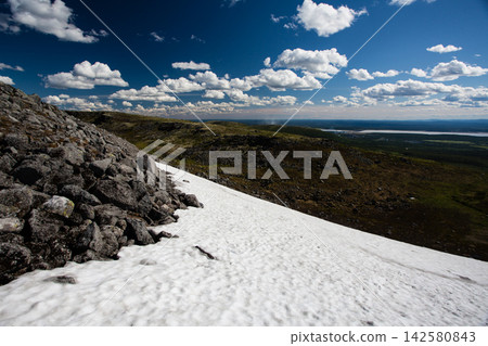 Patch of unmelted snow remaining among dark rocks during summer in the Khibiny Mountains, Murmansk Region, Russia. The lingering mountain snow contrasts with the rugged rocky terrain beneath a deep bl Patch of unmelted snow remaining among dark rocks during summer in the Khibiny Mountains, Murmansk Region, Russia. The lingering mountain snow contrasts with the rugged rocky terrain beneath a deep bl 142580843