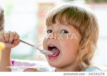 Little girl opens mouth for spoonful of yogurt at breakfast Little girl opens mouth for spoonful of yogurt at breakfast 142581279