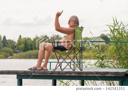 Relaxed senior man raising hand while sitting on lakeside dock Relaxed senior man raising hand while sitting on lakeside dock 142581351