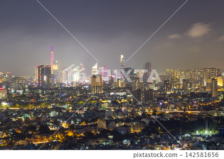A panoramic urban skyline glows with colorful building lights beneath a dark, cloud-filled night sky. 142581656