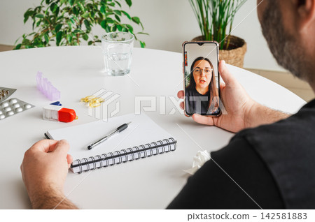 Man Speaking with Doctor on a Video Consultation. Mature man speaks with a female doctor on a smartphone video call at a table with a notebook and pill organizer, illustrating telemedicine, online con Man Speaking with Doctor on a Video Consultation. Mature man speaks with a female doctor on a smartphone video call at a table with a notebook and pill organizer, illustrating telemedicine, online con 142581883
