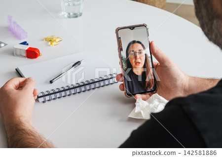 Online Doctor Consultation on Smartphone at Home. Mature man holds a smartphone showing a female doctor during an online consultation at a table with a notebook and tissues, illustrating telemedicine Online Doctor Consultation on Smartphone at Home. Mature man holds a smartphone showing a female doctor during an online consultation at a table with a notebook and tissues, illustrating telemedicine 142581886