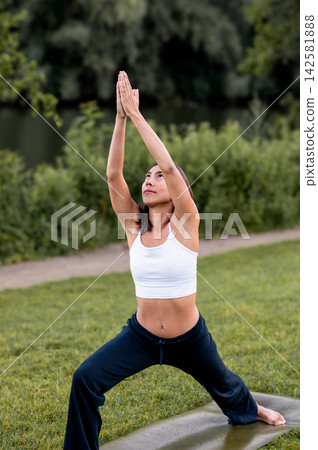 Fit Woman in Crescent Lunge With Prayer Hands on a Yoga Mat Outdoors. A young Hispanic woman practises a crescent lunge with prayer hands on the grass beside the water in the evening light, connecting Fit Woman in Crescent Lunge With Prayer Hands on a Yoga Mat Outdoors. A young Hispanic woman practises a crescent lunge with prayer hands on the grass beside the water in the evening light, connecting 142581888