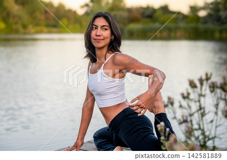 Young Woman in Seated Pose on a Dock by the Lake. A young Hispanic woman practises a seated pose on a wooden dock over the calm lake at sunset, connecting body and mind in nature amid green summer Young Woman in Seated Pose on a Dock by the Lake. A young Hispanic woman practises a seated pose on a wooden dock over the calm lake at sunset, connecting body and mind in nature amid green summer 142581889