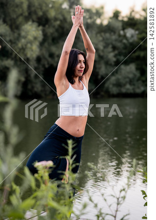 Hispanic Woman in Tree Pose in a Green Park. A young Hispanic woman practises the tree pose in a green park beside a calm lake at sunset, a moment of self care and active summer living at dusk. Hispanic Woman in Tree Pose in a Green Park. A young Hispanic woman practises the tree pose in a green park beside a calm lake at sunset, a moment of self care and active summer living at dusk. 142581892