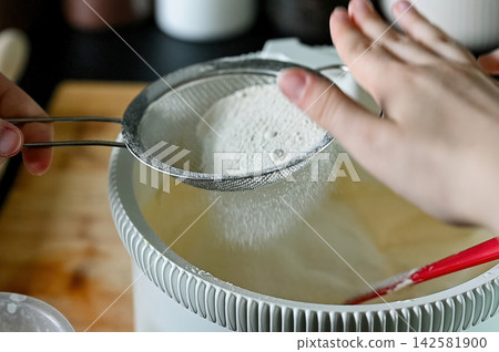 Close-up of Flour Being Sifted into Batter. Macro close-up of fine flour falling through a sieve into a bowl of pale sponge cake batter in a modern kitchen, adding dry ingredients for home baking. 142581900