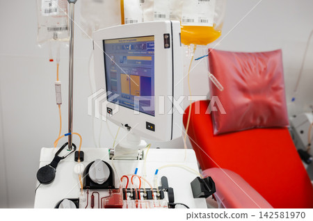 Apheresis Machine and Plasma Bags by Donation Chair, An apheresis machine with a screen and plasma bags on an IV stand beside a red donation chair at a blood bank, illustrating automated plasma collec Apheresis Machine and Plasma Bags by Donation Chair, An apheresis machine with a screen and plasma bags on an IV stand beside a red donation chair at a blood bank, illustrating automated plasma collec 142581970