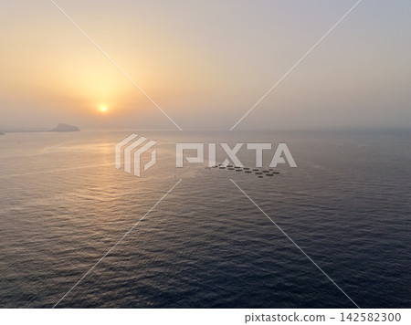 Sunrise over calm sea with distant island silhouette and circular aquaculture pens glowing along horizon under soft pastel sky and warm reflected light, with sunrise, sea, ocean, horizon detail Sunrise over calm sea with distant island silhouette and circular aquaculture pens glowing along horizon under soft pastel sky and warm reflected light, with sunrise, sea, ocean, horizon detail 142582300