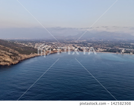 Coastal town and bay from hillside with calm blue sea, rocky headland, clustered shoreline homes, distant mountains and pale sky creating serene wide landscape view Coastal town and bay from hillside with calm blue sea, rocky headland, clustered shoreline homes, distant mountains and pale sky creating serene wide landscape view 142582308