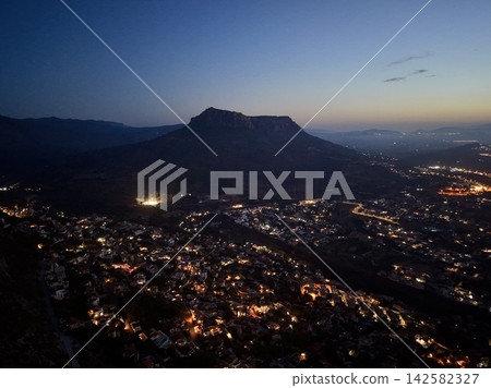 Silhouetted mesa above glowing coastal, town at dusk with deep blue sky, distant hills, warm streetlights, layered horizon, and calm evening atmosphere in wide aerial view Silhouetted mesa above glowing coastal, town at dusk with deep blue sky, distant hills, warm streetlights, layered horizon, and calm evening atmosphere in wide aerial view 142582327