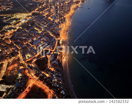 Twilight aerial view of illuminated beachfront city with curving coastline, high-rise towers, glowing promenade, sandy shore, and calm dark sea, with beachfront, coastline, cityscape, aerial detail Twilight aerial view of illuminated beachfront city with curving coastline, high-rise towers, glowing promenade, sandy shore, and calm dark sea, with beachfront, coastline, cityscape, aerial detail 142582332