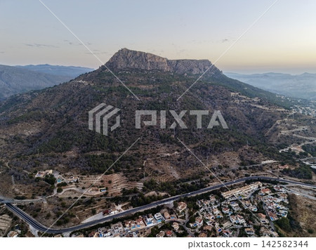 Rocky plateau rising above terraced pine hills, winding roads, seaside homes, and coastal town under soft dawn sky with pastel light and calm distant horizon, with mountain, plateau, cliff, rocky Rocky plateau rising above terraced pine hills, winding roads, seaside homes, and coastal town under soft dawn sky with pastel light and calm distant horizon, with mountain, plateau, cliff, rocky 142582344