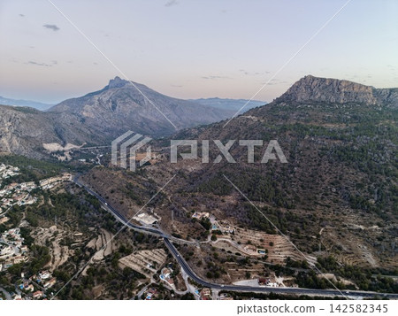 Dusk mountain valley with winding road, scattered houses, rocky ridges, terraced slopes, pine woodland, and soft pastel sky across wide scenic landscape, with mountain, valley, peak, ridge detail Dusk mountain valley with winding road, scattered houses, rocky ridges, terraced slopes, pine woodland, and soft pastel sky across wide scenic landscape, with mountain, valley, peak, ridge detail 142582345