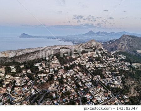 aerial view of a coastal town with residential buildings, pools, and roads amidst mountainous terrain and calm waters, peaceful and serene perspective from above. aerial view of a coastal town with residential buildings, pools, and roads amidst mountainous terrain and calm waters, peaceful and serene perspective from above. 142582347