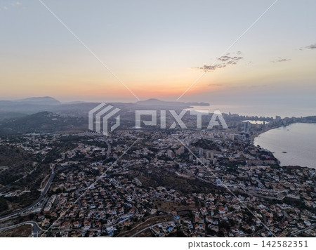 Aerial coastal cityscape at pastel sunrise with misty mountains, calm sea, shoreline neighborhoods, bayfront highrises, and soft morning haze over scenic urban waterfront Aerial coastal cityscape at pastel sunrise with misty mountains, calm sea, shoreline neighborhoods, bayfront highrises, and soft morning haze over scenic urban waterfront 142582351