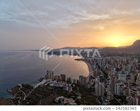 Aerial coastal city skyline beside curved sandy beach at sunset, distant mountains, calm sea, warm light, and urban waterfront creating scenic travel landscape, with sunset, coast, city, beach detail Aerial coastal city skyline beside curved sandy beach at sunset, distant mountains, calm sea, warm light, and urban waterfront creating scenic travel landscape, with sunset, coast, city, beach detail 142582365