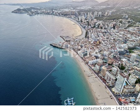 Aerial view of coastal city with curved sandy bay, highrise buildings, turquoise sea, marina, promenade, and mountain backdrop at dusk, with coast, bay, beach, sea detail Aerial view of coastal city with curved sandy bay, highrise buildings, turquoise sea, marina, promenade, and mountain backdrop at dusk, with coast, bay, beach, sea detail 142582373