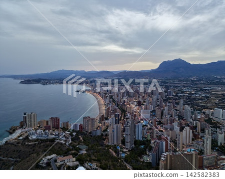 Coastal city skyline at dusk with curved sandy beach, calm sea, mountain backdrop, rocky headland, glowing streetlights, dense high-rise district, overcast sky and wide waterfront panorama 142582383