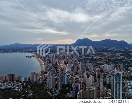 Coastal city skyline at dusk with curved beachfront, glowing promenade, high rise towers, calm bay water, and mountain silhouette beneath cloudy evening sky, with city, coast, beach, bay detail Coastal city skyline at dusk with curved beachfront, glowing promenade, high rise towers, calm bay water, and mountain silhouette beneath cloudy evening sky, with city, coast, beach, bay detail 142582384