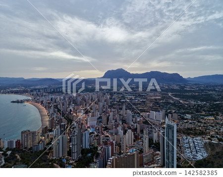 Coastal city skyline at dusk with mountain backdrop, illuminated waterfront, curved sandy bay, urban district, and dramatic cloudscape over calm seascape, with city, coastal, skyline, dusk detail Coastal city skyline at dusk with mountain backdrop, illuminated waterfront, curved sandy bay, urban district, and dramatic cloudscape over calm seascape, with city, coastal, skyline, dusk detail 142582385