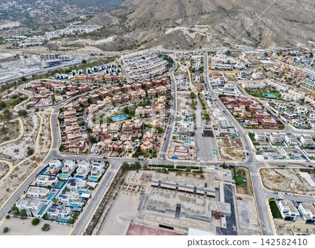 High angled aerial view showing dense coastal hillside residential neighborhood with terracotta roofs, white villas, winding roads, pools, mountain backdrop, sunlit urban layout High angled aerial view showing dense coastal hillside residential neighborhood with terracotta roofs, white villas, winding roads, pools, mountain backdrop, sunlit urban layout 142582410