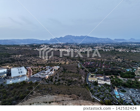 Coastal town spread across rolling hills beneath pale blue dusk sky, with resort pools, industrial foreground, and distant mountain ridge in panoramic aerial view Coastal town spread across rolling hills beneath pale blue dusk sky, with resort pools, industrial foreground, and distant mountain ridge in panoramic aerial view 142582424