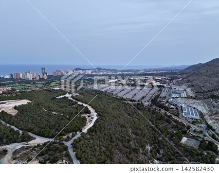 Wide aerial view of coastal city skyline with highrise towers, forested hillside, resort district, roads, parking areas and calm sea under pale blue sky in distance Wide aerial view of coastal city skyline with highrise towers, forested hillside, resort district, roads, parking areas and calm sea under pale blue sky in distance 142582430