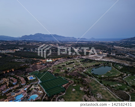 Coastal town panorama with mountain backdrop at dusk, illuminated resort pools, golf fairways, lakes, sports fields, winding roads, residential areas, shoreline, bay and surrounding foothills Coastal town panorama with mountain backdrop at dusk, illuminated resort pools, golf fairways, lakes, sports fields, winding roads, residential areas, shoreline, bay and surrounding foothills 142582431