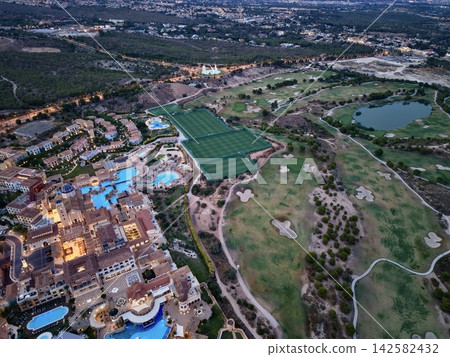 Aerial view of resort pools, sports fields, and adjacent golf course at dusk with illuminated pathways, hotel buildings, residential cluster, and landscaped grounds Aerial view of resort pools, sports fields, and adjacent golf course at dusk with illuminated pathways, hotel buildings, residential cluster, and landscaped grounds 142582432