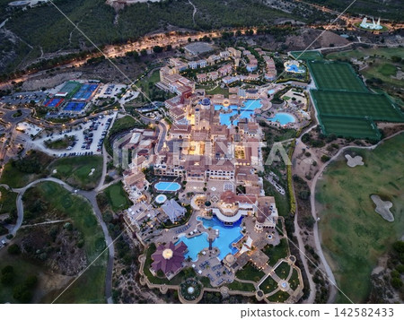 High angle evening view of illuminated resort complex with pools, courts, parking areas, soccer fields, terraces, and wooded hills beside Mediterranean style landscape High angle evening view of illuminated resort complex with pools, courts, parking areas, soccer fields, terraces, and wooded hills beside Mediterranean style landscape 142582433