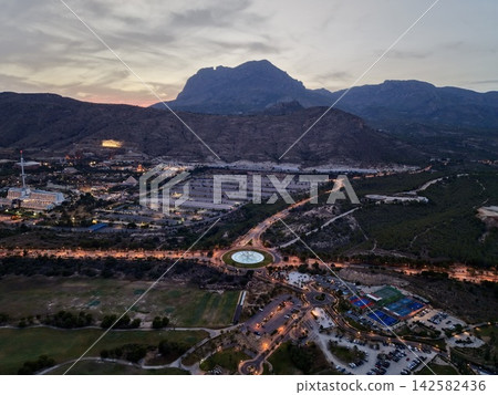 Sunset aerial panorama of coastal resort town with glowing roundabout, fountain sculpture, hotel district, roads, and mountain silhouette rising over shoreline, with mountain, resort, aerial, dusk Sunset aerial panorama of coastal resort town with glowing roundabout, fountain sculpture, hotel district, roads, and mountain silhouette rising over shoreline, with mountain, resort, aerial, dusk 142582436