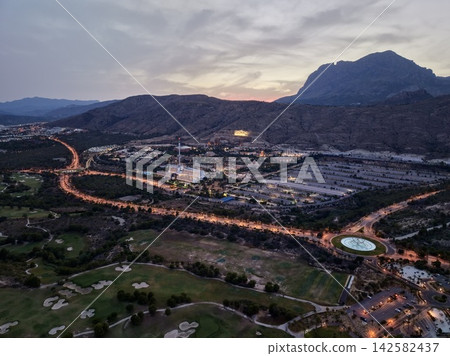 Dusk aerial view of coastal resort town below rugged mountains with glowing roads, golf course greens, hillside homes, shoreline atmosphere, and wide scenic urban landscape Dusk aerial view of coastal resort town below rugged mountains with glowing roads, golf course greens, hillside homes, shoreline atmosphere, and wide scenic urban landscape 142582437