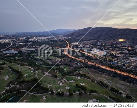 Aerial view of coastal town at dusk with illuminated roads, golf course, hotel complex, amusement rides, and cloudy sky over distant water and hills, with aerial, coastal, town, dusk detail Aerial view of coastal town at dusk with illuminated roads, golf course, hotel complex, amusement rides, and cloudy sky over distant water and hills, with aerial, coastal, town, dusk detail 142582438