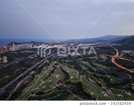 Coastal city skyline at evening beside illuminated highway, green golf course, winding shoreline, calm sea and distant mountains under soft dusk light, with aerial, coastal, city, skyline detail Coastal city skyline at evening beside illuminated highway, green golf course, winding shoreline, calm sea and distant mountains under soft dusk light, with aerial, coastal, city, skyline detail 142582439